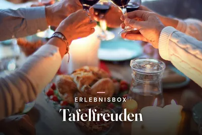 Friends raising glasses of red wine over a candlelit dinner table, with the text "Erlebnisbox Tafelfreuden"