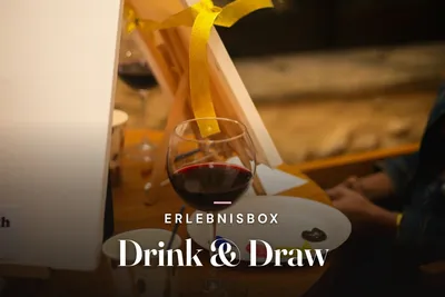 Glass of red wine next to a small easel, paint palette and brushes in warm evening light, with the title "Erlebnisbox Drink & Draw".