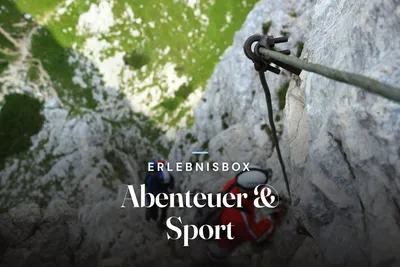 View down from a via ferrata: a steel safety cable bolted to a limestone rock face, with two climbers in helmets below and green meadows far beneath, with the text “Erlebnisbox – Abenteuer & Sport”