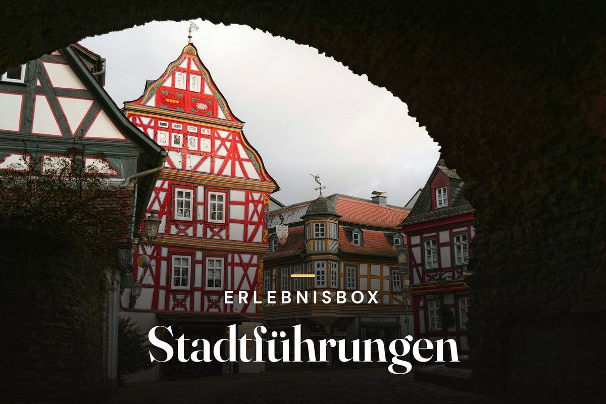 Colourful red-and-white half-timbered houses in a historic old town, seen through a dark stone archway, with the text “Erlebnisbox – Stadtführungen”