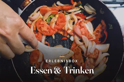 Hand stirring sliced onions, tomatoes and peppers in a frying pan, seen from above, with the text “Erlebnisbox – Essen & Trinken”