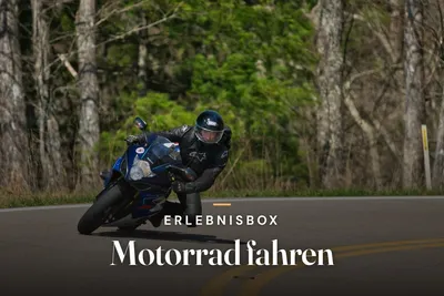 Motorcyclist in black leathers leaning a blue sports bike into a curve on a country road lined with trees, with the text “Erlebnisbox – Motorrad fahren”