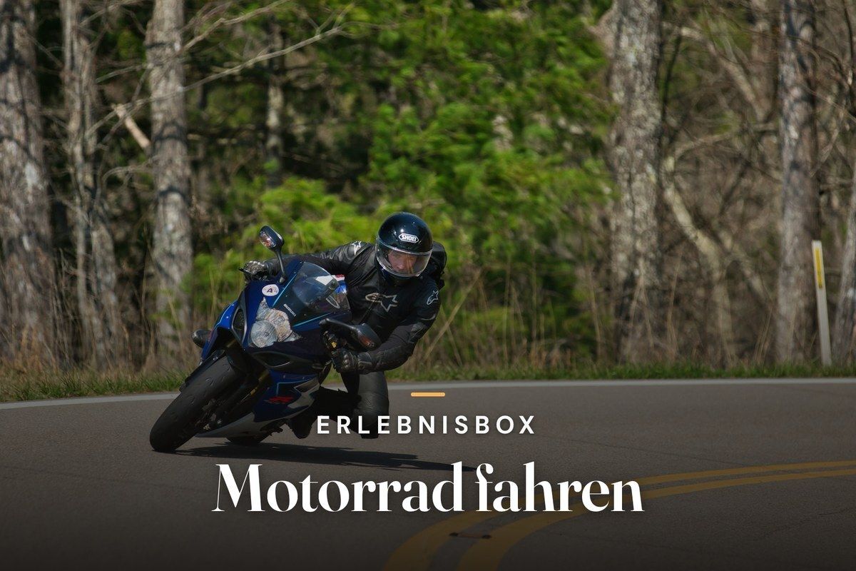 Motorcyclist in black leathers leaning a blue sports bike into a curve on a country road lined with trees, with the text “Erlebnisbox – Motorrad fahren”