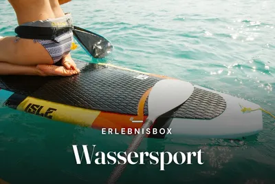 Person in a life belt kneeling on a stand-up paddleboard with a paddle on turquoise, sunlit water, with the text “Erlebnisbox – Wassersport”