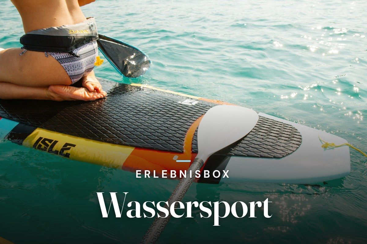 Person in a life belt kneeling on a stand-up paddleboard with a paddle on turquoise, sunlit water, with the text “Erlebnisbox – Wassersport”