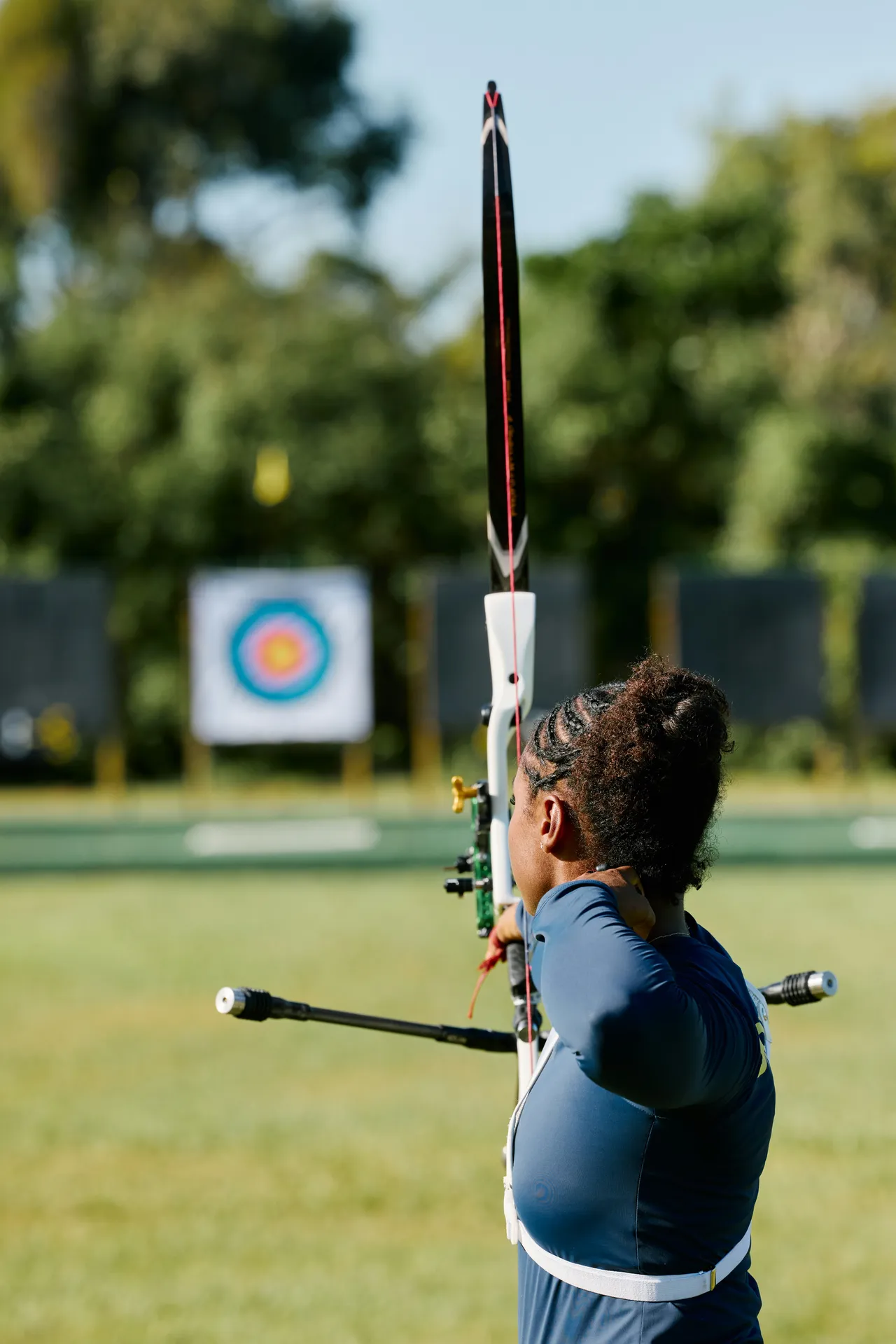 Back view of a female archer with braided hair pulled into a bun, aiming a modern recurve bow at a distant bullseye target on an outdoor range.