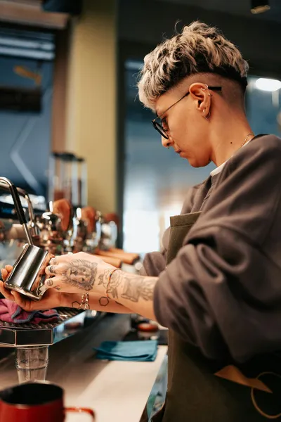 Tattooed female barista with short hair and glasses working at an espresso machine, focused on preparing or adjusting the equipment.