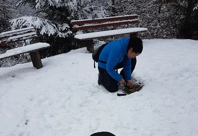Person in blue jacket crouching in snow with wooden benches trying to light a fire