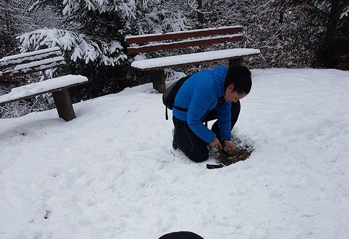 Person in blue jacket crouching in snow with wooden benches trying to light a fire