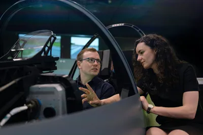 An instructor standing beside a person using a virtual reality flight simulator, guiding them through the training setup.