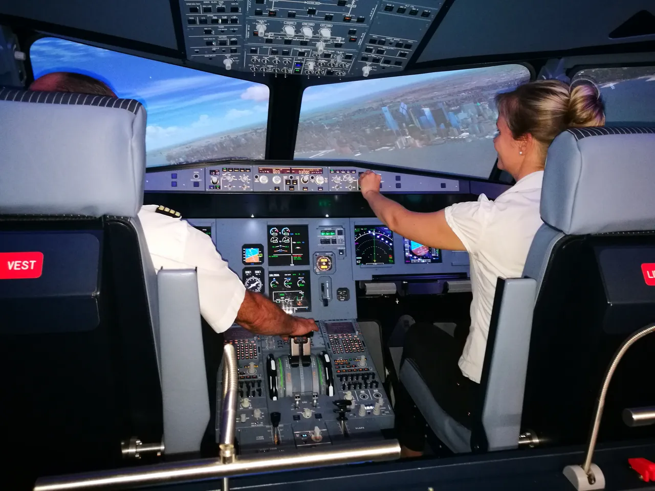Two people in the flight simulator cockpit approaching a New York City skyline, with the woman on the right adjusting the controls.