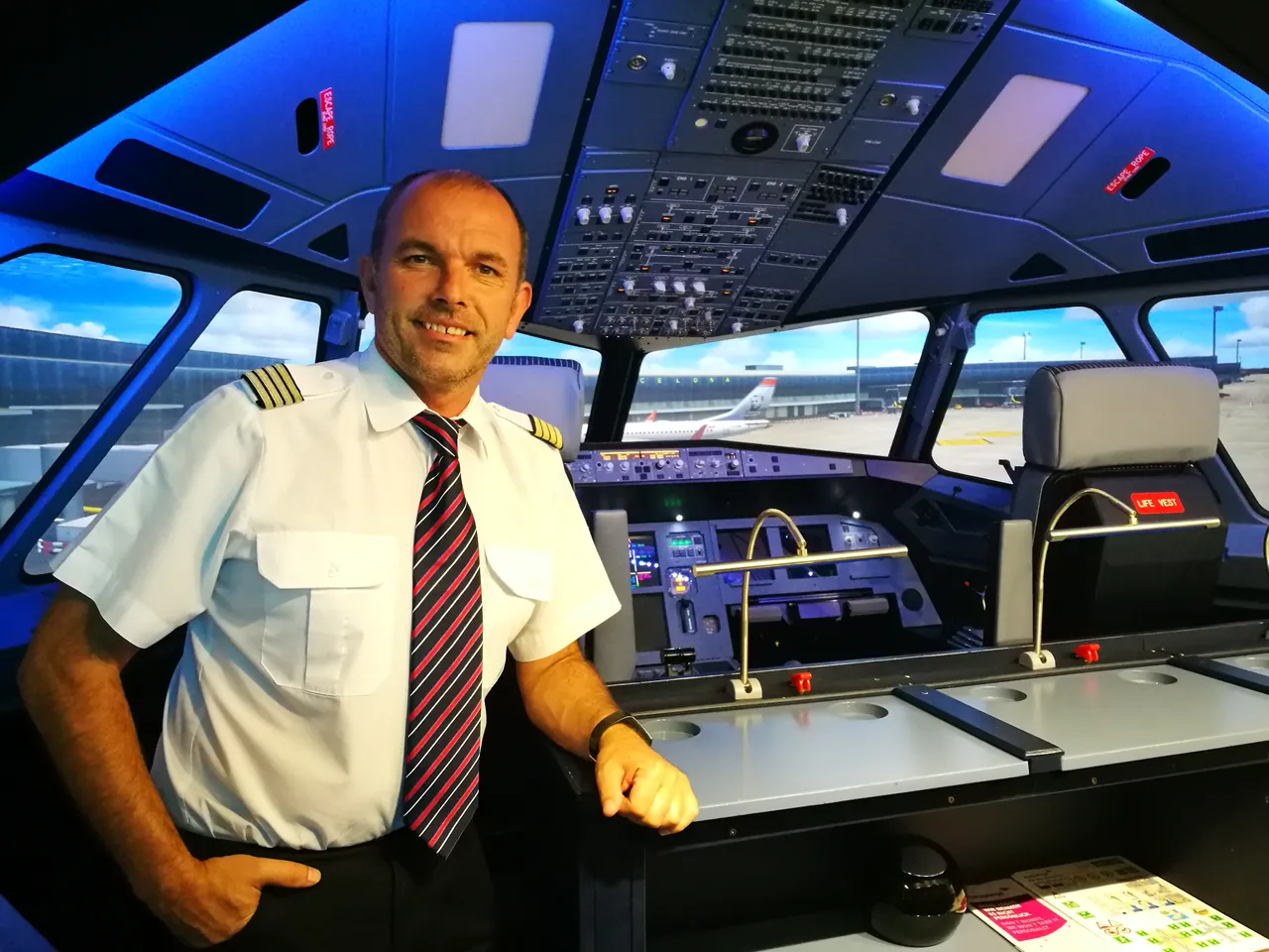 Man in pilot uniform standing at the front of the flight simulator cockpit, with a view of Barcelona airport and an aircraft in the background.