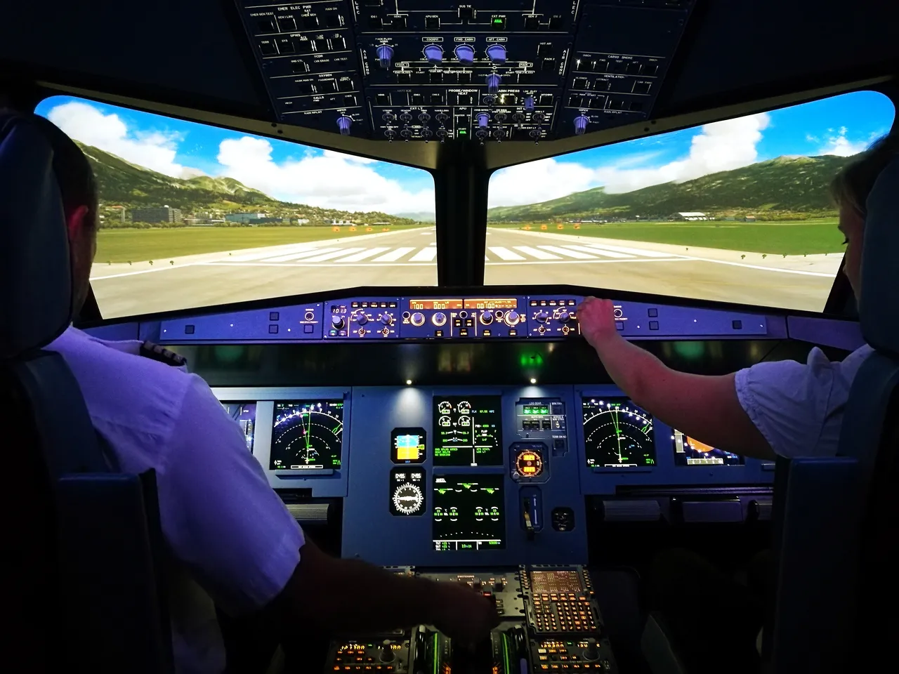 View from inside the flight simulator cockpit of two people preparing to land on a runway surrounded by green mountains.