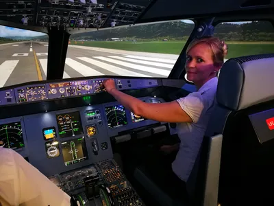 Woman seated at the flight simulator controls, smiling at the camera with a runway and green hills visible through the cockpit windows.