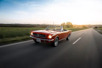 Red 1965 Ford Mustang convertible driving on a country road at sunset
