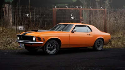 Orange 1970 Ford Mustang with black stripes in front of an old industrial site