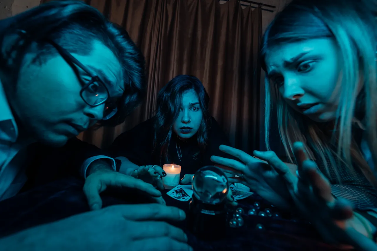 Three people with shocked and intense expressions looking down at a glowing crystal ball and playing cards under dramatic blue lighting.