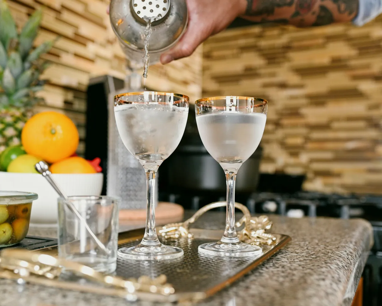 A barista pouring gin from a shaker into a glass, on a bar with fruits for cocktails behind it.