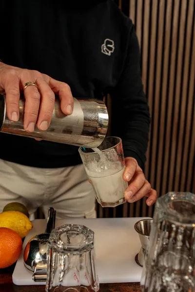 Close-up of hands pouring a gin and tonic from a shaker into a glass, with citrus fruits on the cutting board.