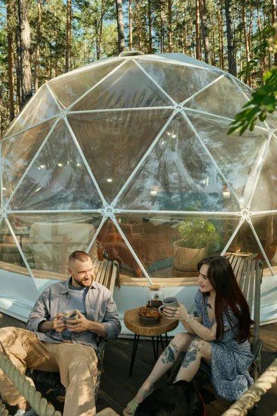 A couple relaxes on wooden chairs outside a transparent geodesic glamping dome in a forest, enjoying drinks and chatting on a sunny deck.