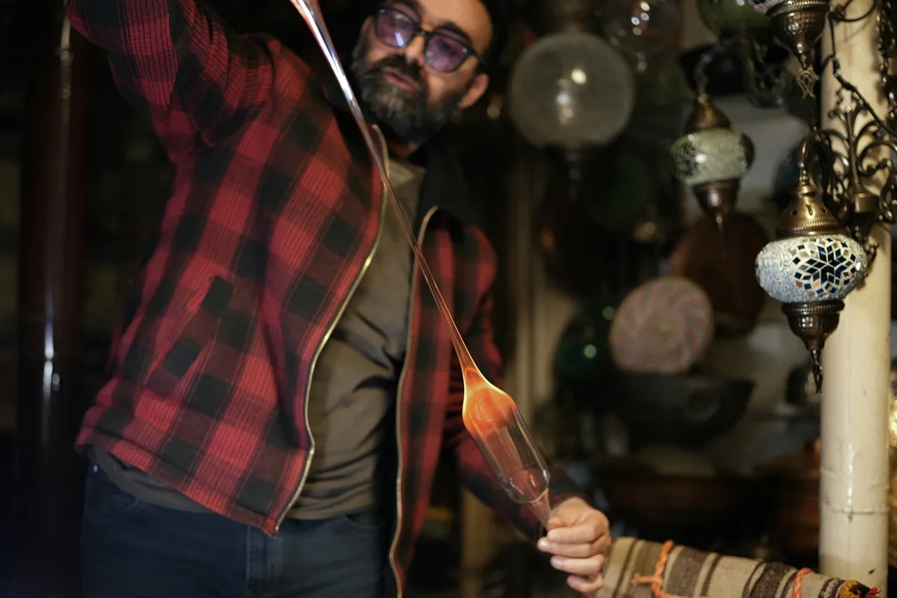 Man shaping a glowing molten glass bulb on a rod, decorative lamps in the background
