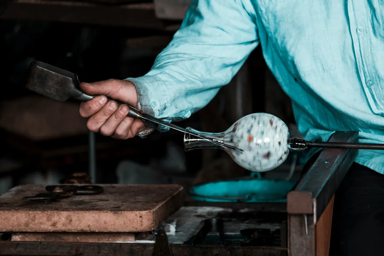 Glassblower using a metal tool to shape a speckled bulbous glass vase on a blowpipe
