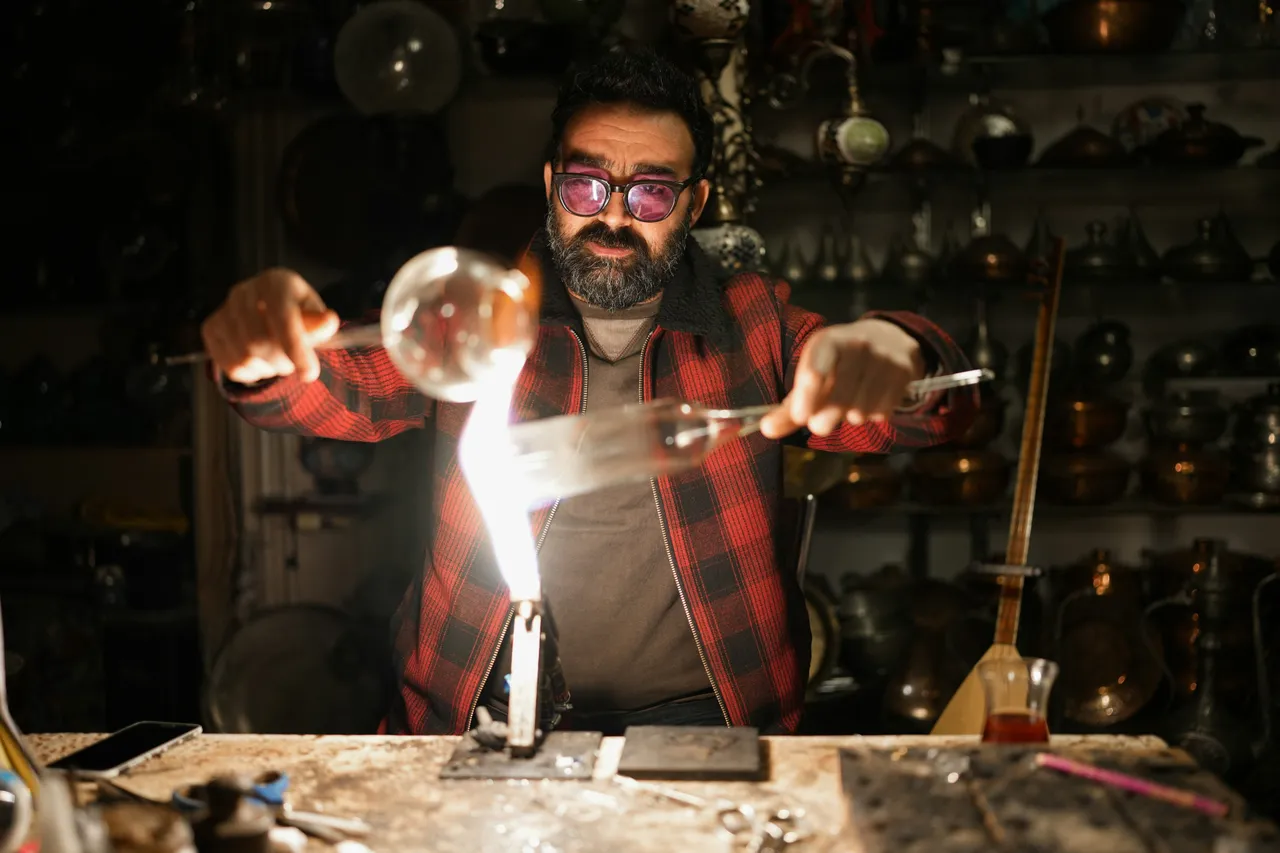 Bearded glassblower in tinted safety glasses shaping molten glass with tools in a workshop
