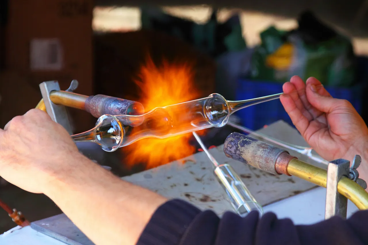 Glassblower's hands shaping a clear glass tube over an open torch flame