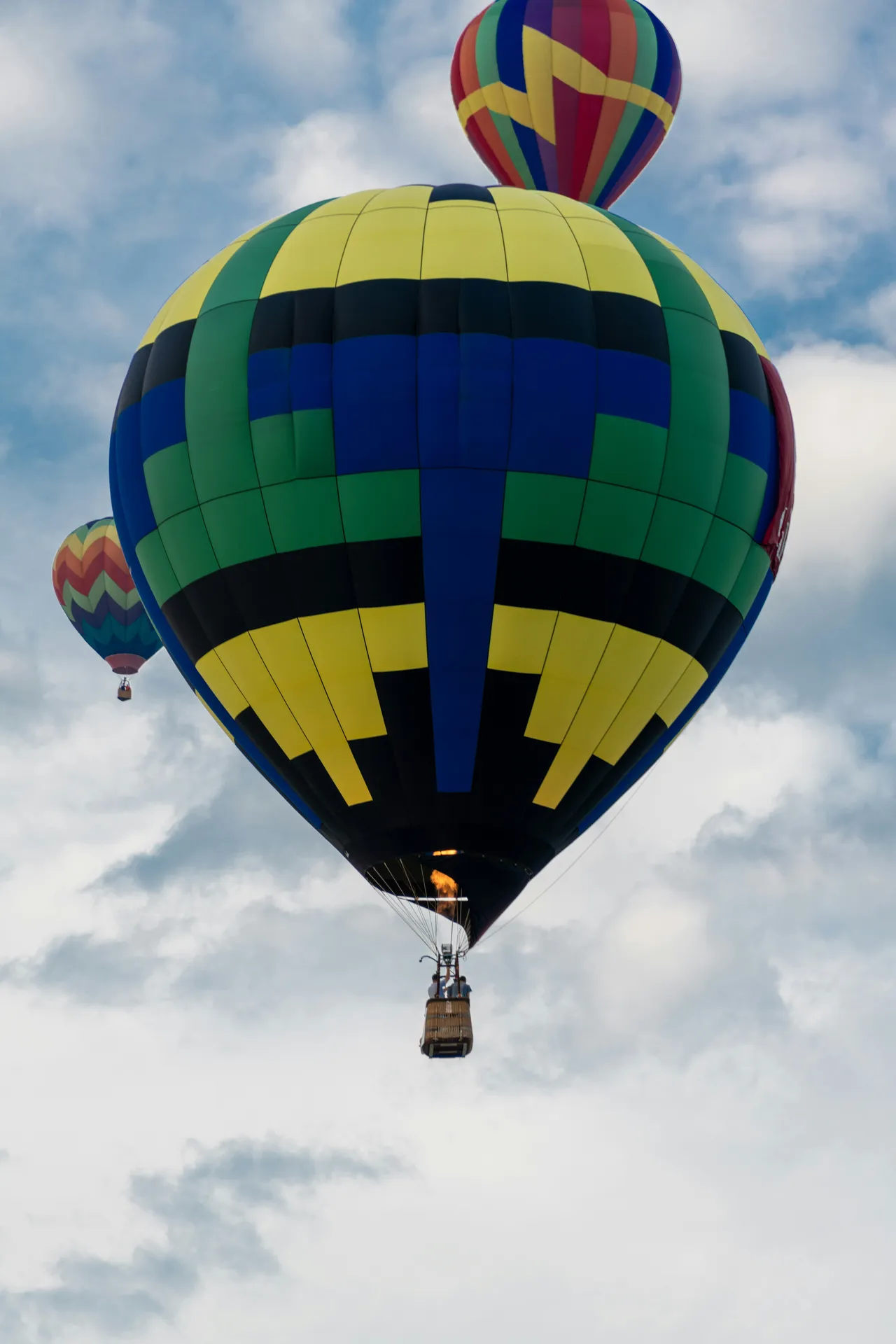 A large green, blue, yellow, and black geometric patterned hot air balloon flies with its burner lit, while two other colorful balloons drift against a cloudy sky.