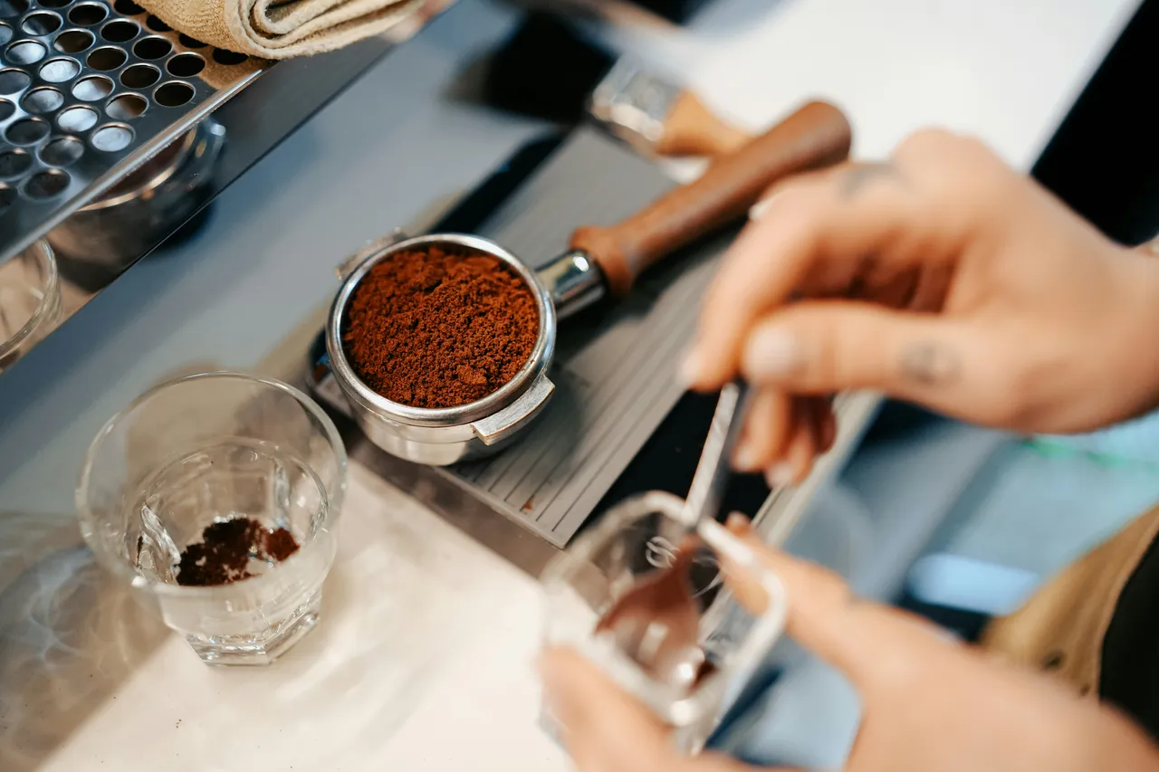"Close-up of finely ground espresso coffee in a metal portafilter basket, with a tamper, towel, and water glass visible on the work surface.