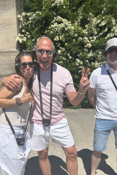 Smiling guests wearing wireless headphones pose in front of a flowering hedge on a sunny day