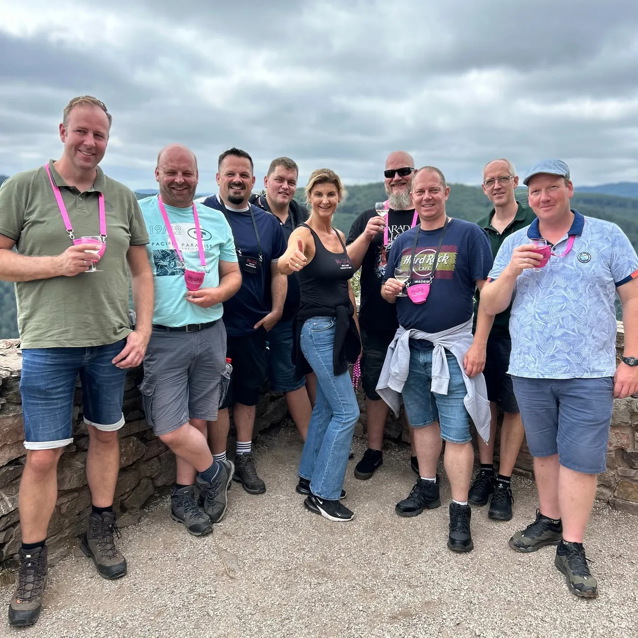 Smiling group of hikers with wine glasses on neck lanyards and their guide giving a thumbs up at a hilltop viewpoint over forested hills.