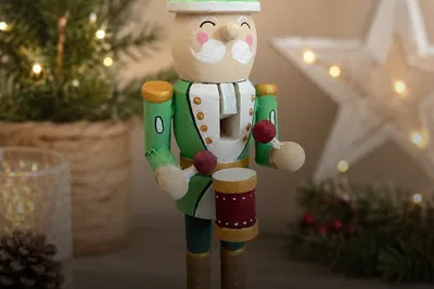 Hand-painted wooden nutcracker with a white moustache, green uniform and red drum, displayed among festive Christmas decorations with fairy lights.