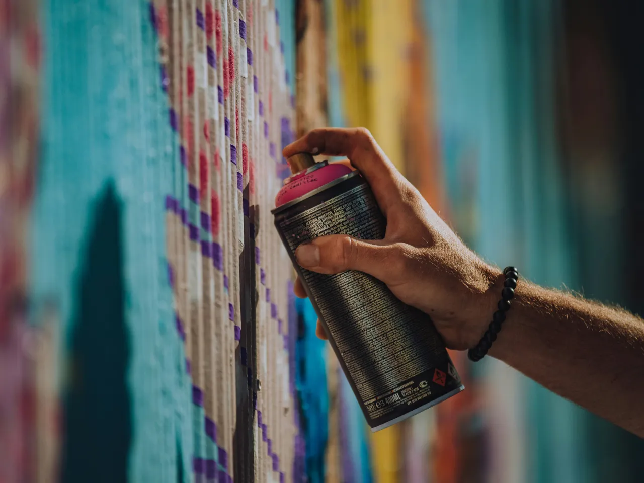 Hand holding a pink spray paint can against a colorful graffiti mural wall
