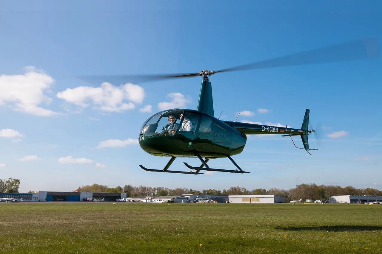 Dark green Robinson R44 helicopter hovering above an airfield under a blue sky