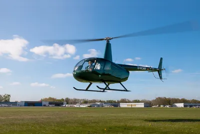 Dark green Robinson R44 helicopter hovering above an airfield under a blue sky
