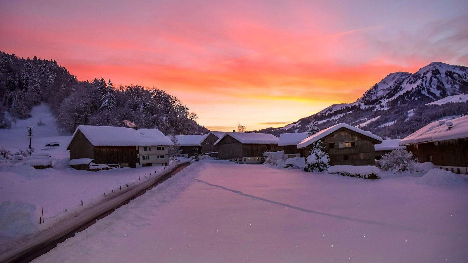 Snow-covered alpine village with wooden farmhouses and a cleared road at sunset, pink and orange sky over snowy mountains and forest