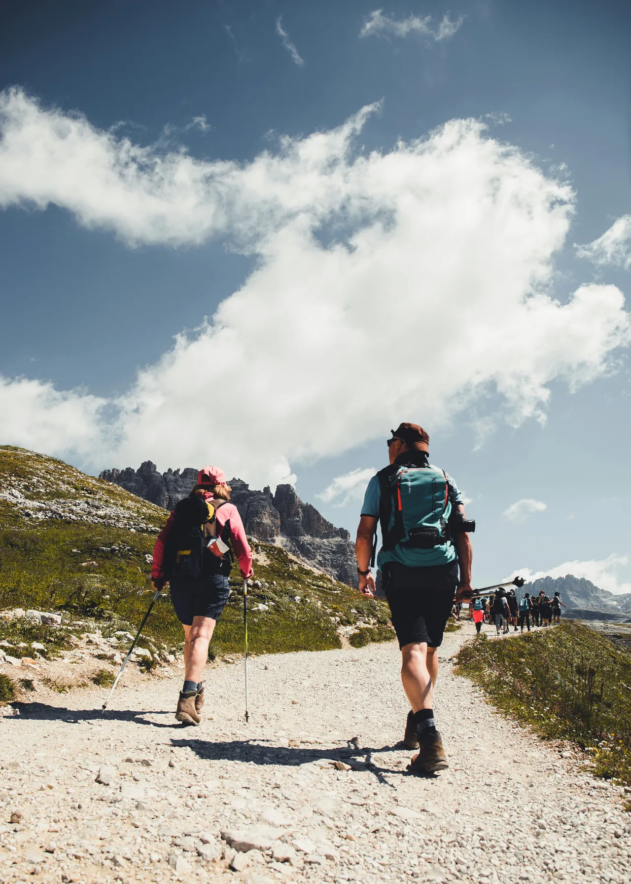 Rear view of a man and a woman with backpacks and trekking poles hiking up a rocky gravel trail toward rugged mountain peaks under a blue sky with fluffy white clouds.