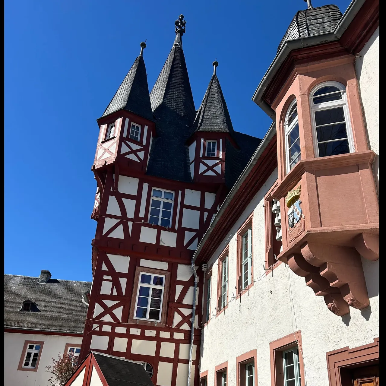 Historic red and white half-timbered building with three pointed slate spires and a sandstone bay window against a blue sky.