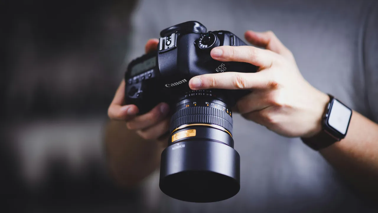 Close-up of a person's hands holding a black Canon DSLR camera equipped with a large prime lens and lens hood.