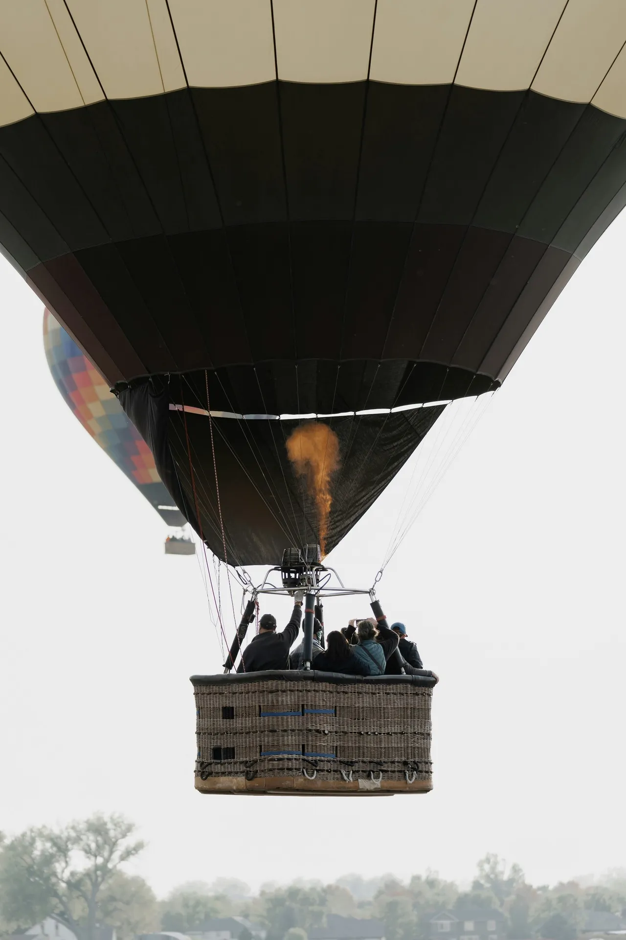 Passengers riding in the basket of a dark hot air balloon with the burner firing, flying high above a hazy landscape.