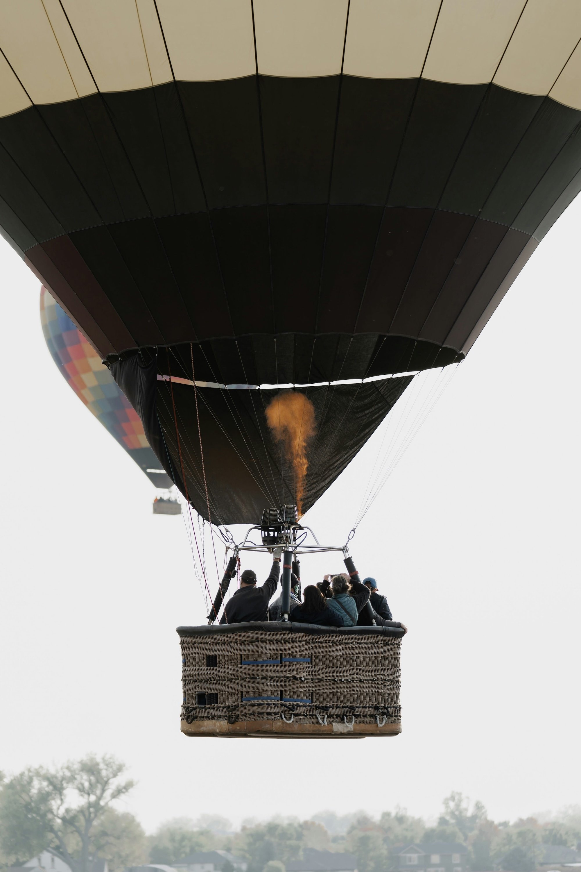 Passengers riding in the basket of a dark hot air balloon with the burner firing, flying high above a hazy landscape.
