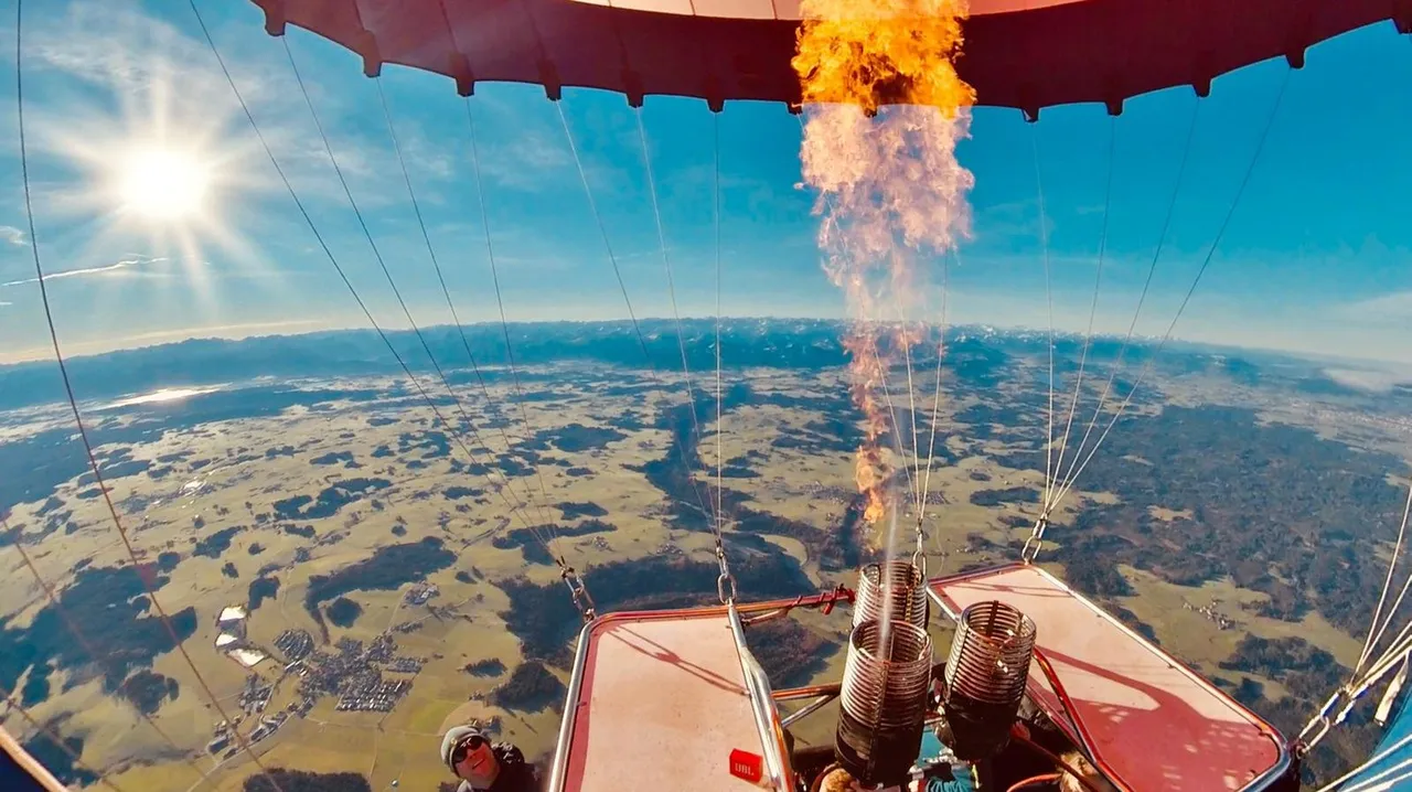 A wide-angle view from inside a hot air balloon basket showing the burner firing flame against a sunny sky and snow-dusted ground below.