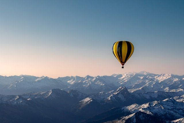 A black and yellow hot air balloon flying high above a snow-covered mountain range under a clear sky.
