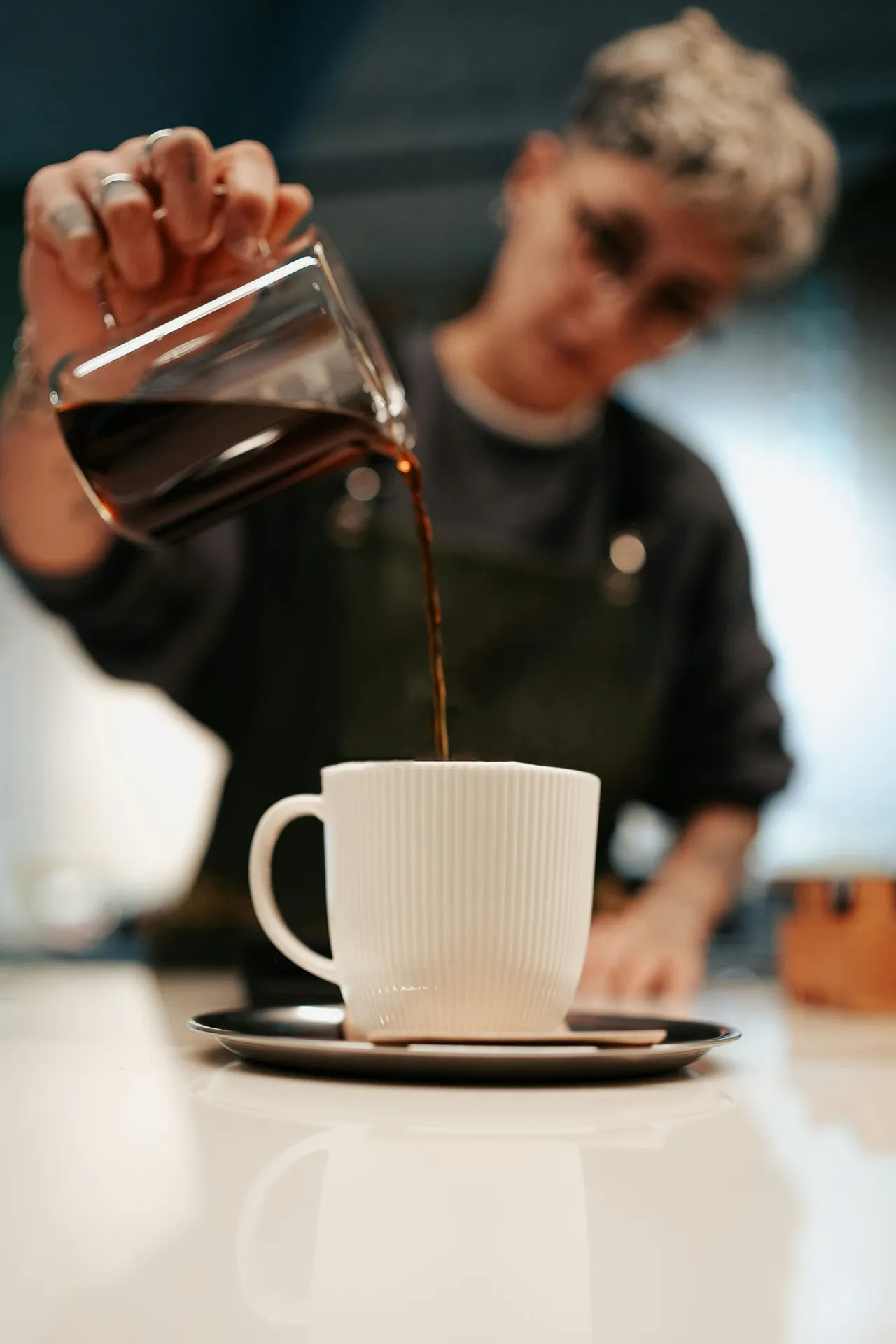 A glass of iced coffee with visible milk layers stands on a dark surface next to a glass bowl filled with whole roasted coffee beans.