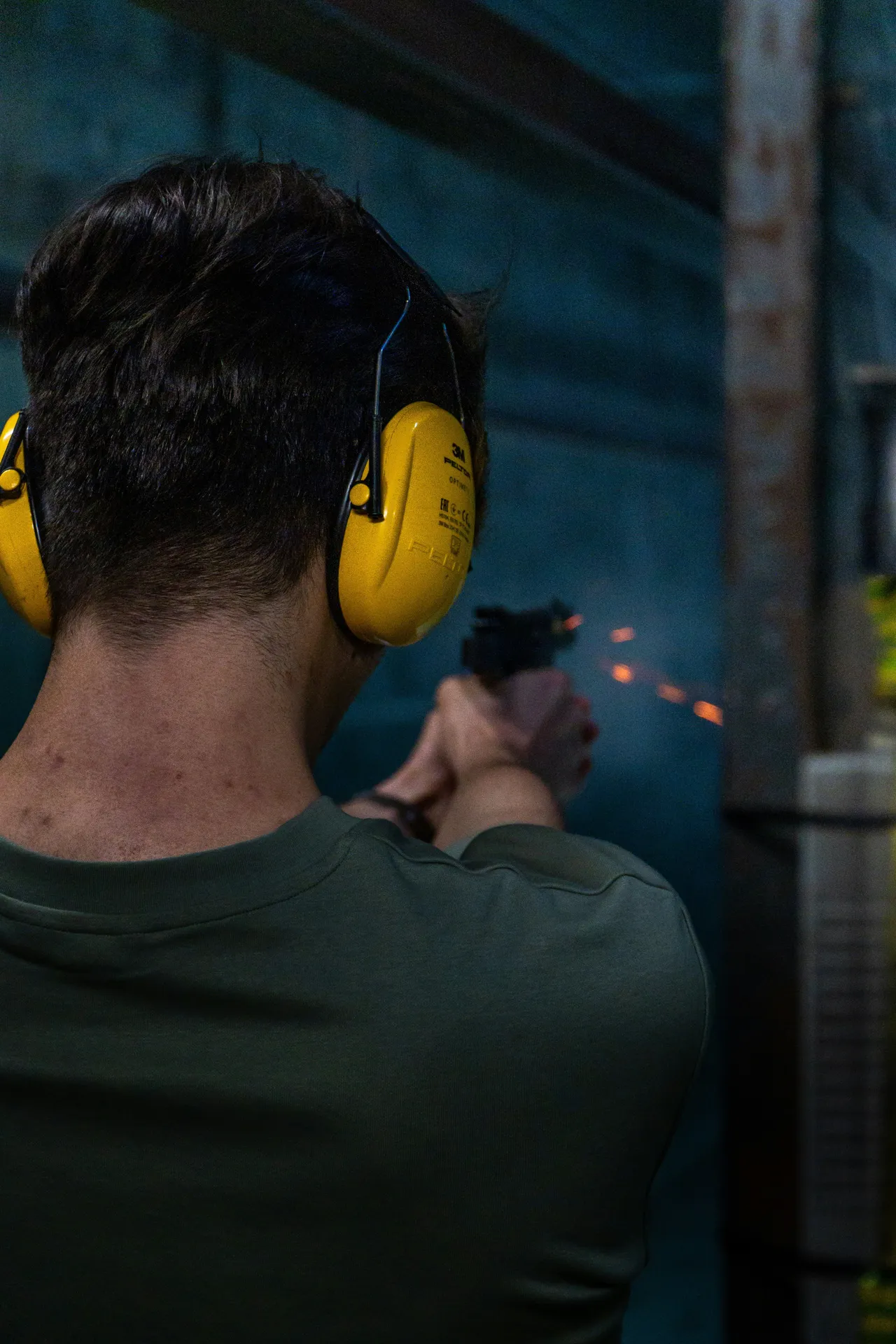 Rear view of a person wearing yellow ear protection firing a handgun at an indoor shooting range, with visible sparks from the muzzle flash.