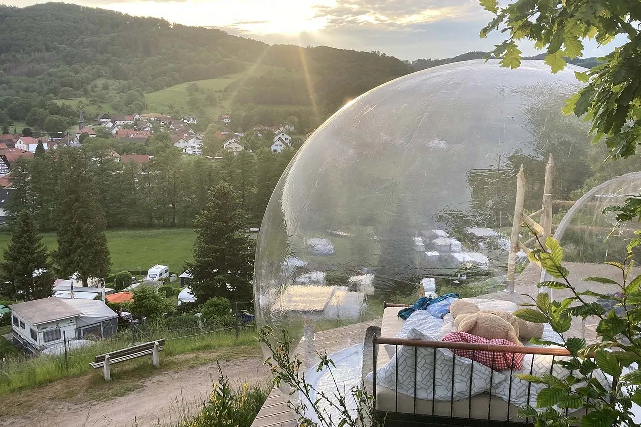 Inflatable transparent bubble tent with a bed on a hillside above a campsite at sunset