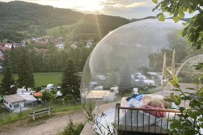 Inflatable transparent bubble tent with a bed on a hillside above a campsite at sunset