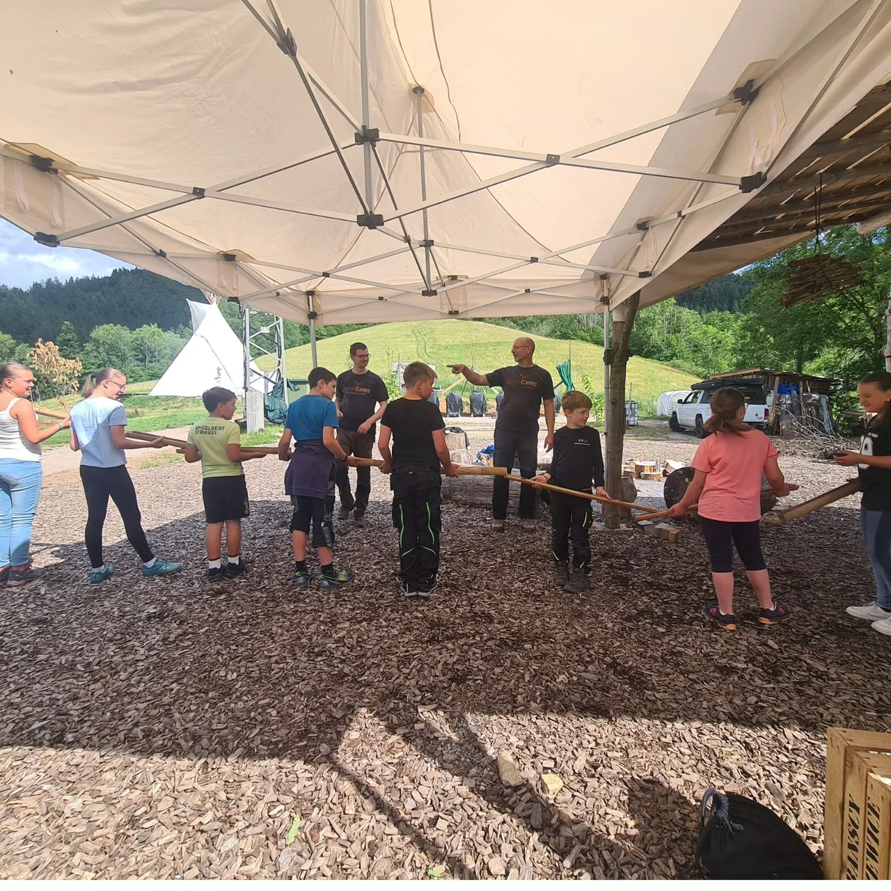 A group of children holding a long wooden pole together during a teamwork exercise under a large white canopy tent at a camp.