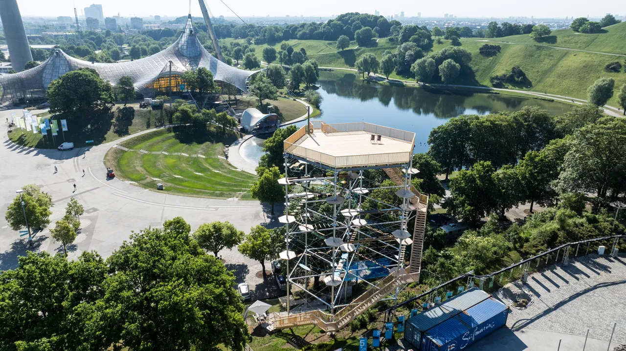 An aerial view of the multi-level Kristallturm high ropes climbing structure with a viewing platform, located next to the lake and distinctive tent-style roofs at Olympiapark in Munich.